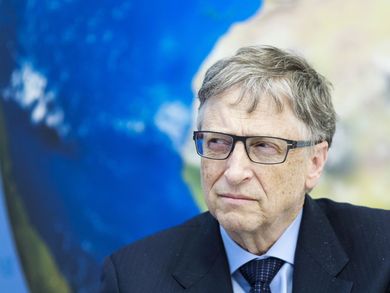 Bill Gates squints while sitting in front of a picture of Earth.