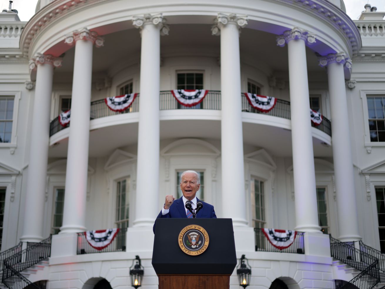 President Biden speaks from behind a lectern and in front of the White House, which is festooned with red, white, and blue bunting.